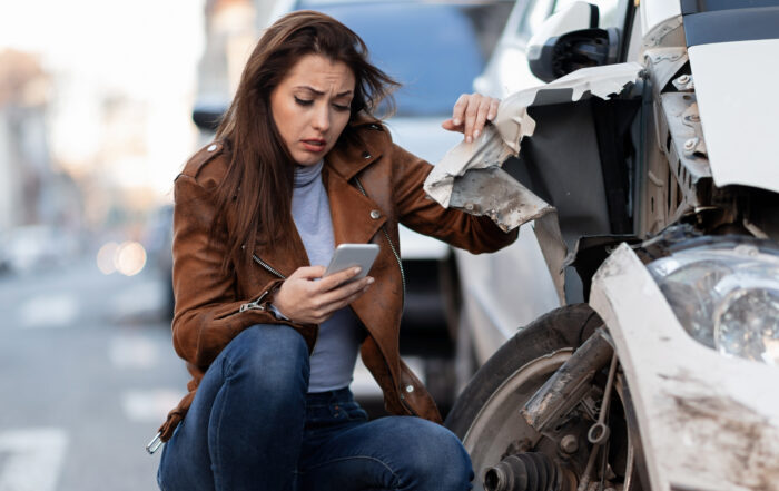 Mujer frente a un auto dañado tras un accidente de tránsito, revisa su celular en busca de asistencia dentro del mercado asegurador.
