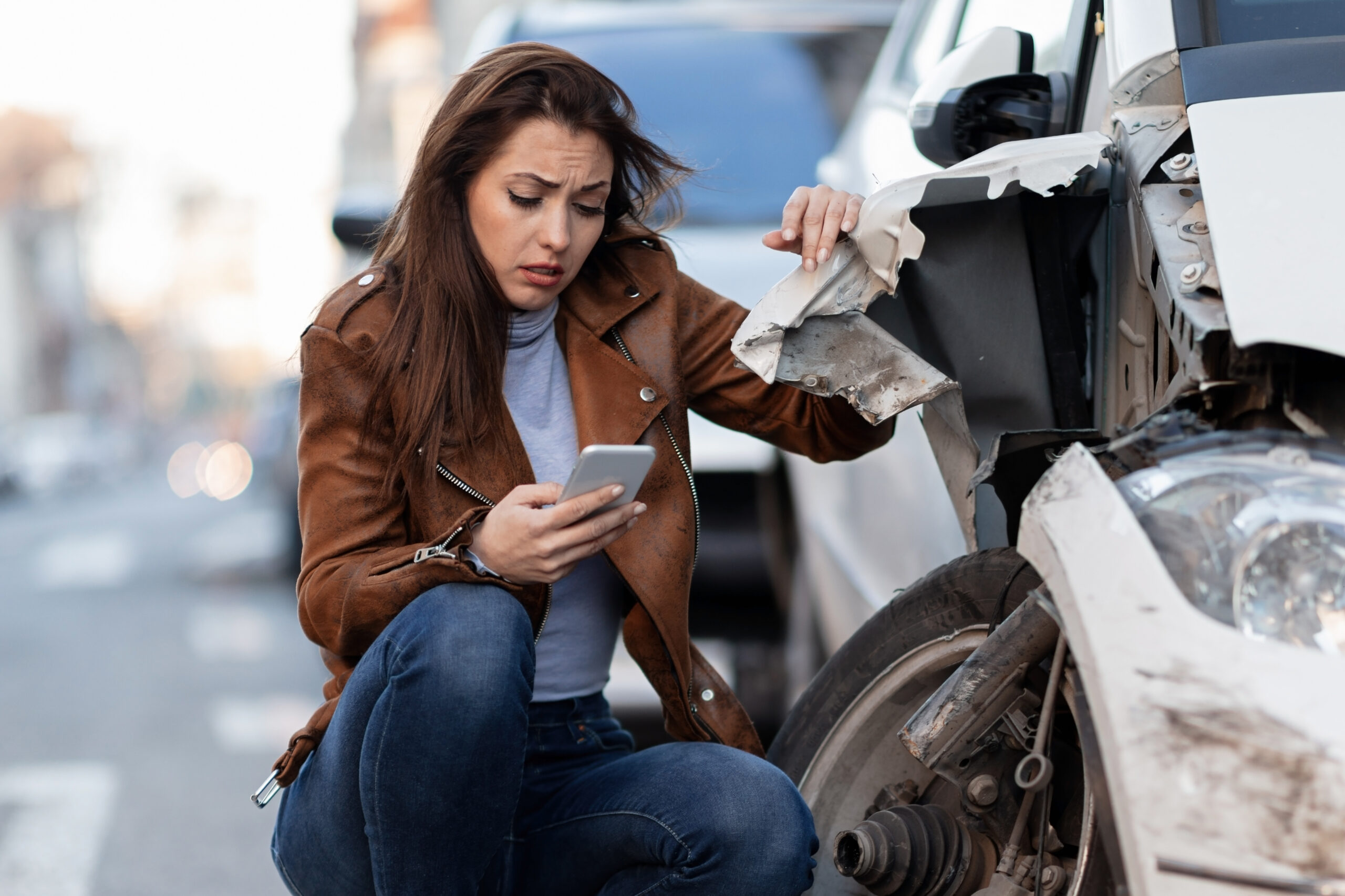 Mujer frente a un auto dañado tras un accidente de tránsito, revisa su celular en busca de asistencia dentro del mercado asegurador.