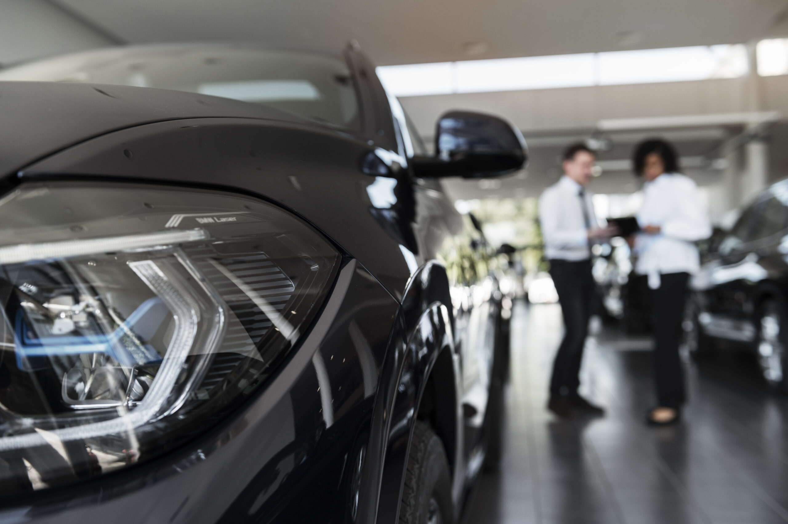 Primer plano de un auto negro en concesionaria con dos personas conversando al fondo sobre opciones de compra de auto.