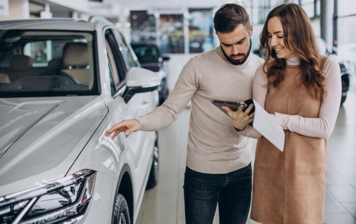 Pareja observa un auto blanco en una concesionaria mientras revisa información en una tablet, proceso común antes de adquirir un auto.
