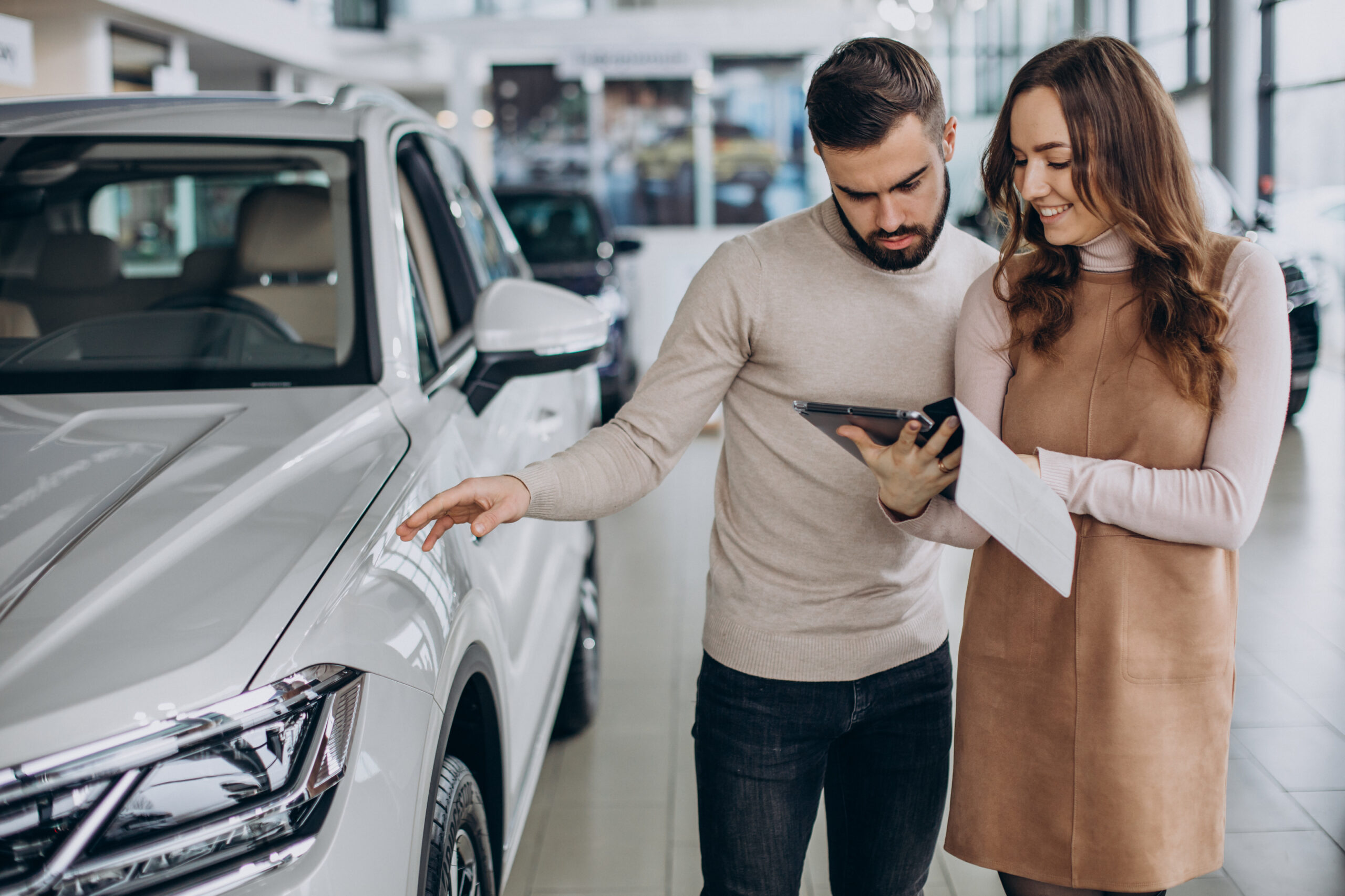 Pareja observa un auto blanco en una concesionaria mientras revisa información en una tablet, proceso común antes de adquirir un auto.